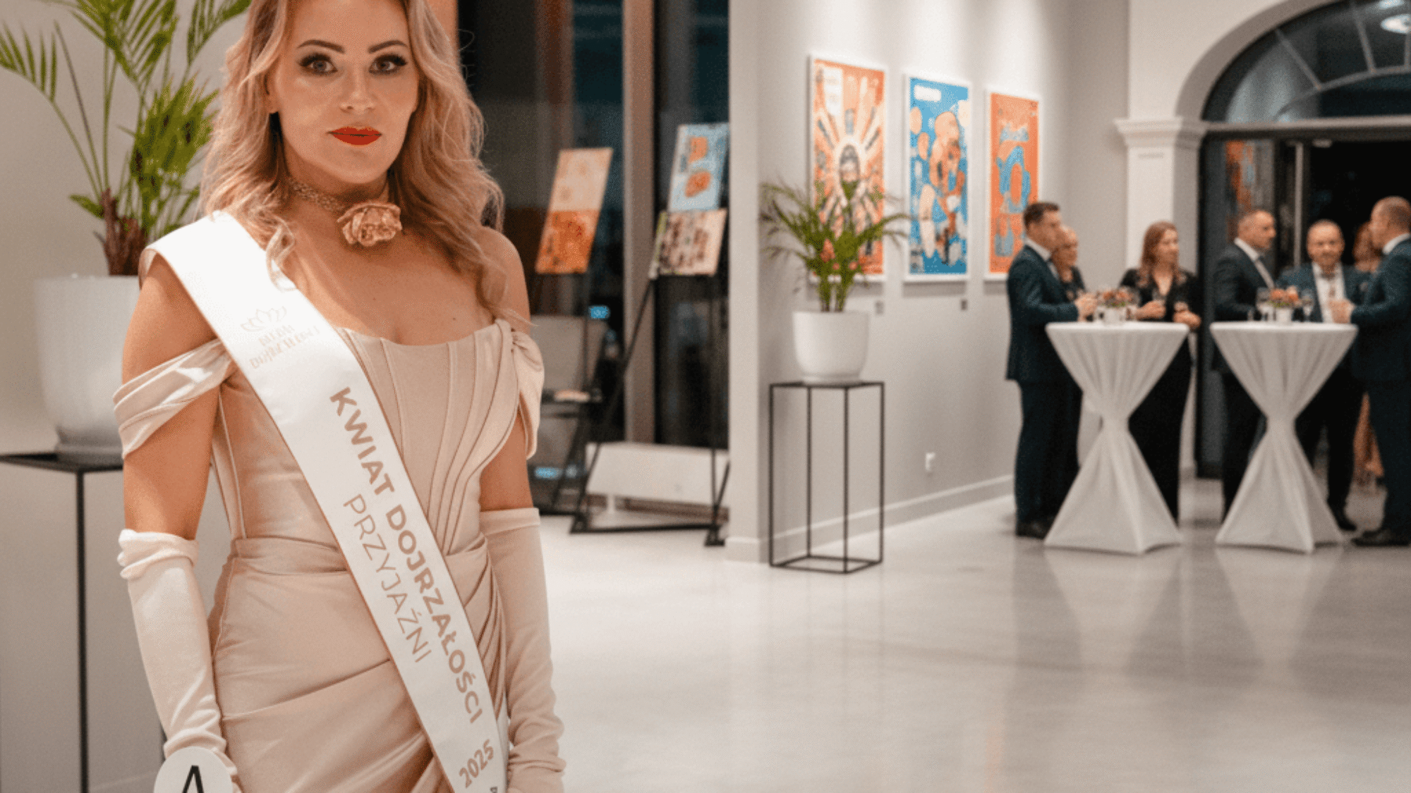 Izabela Kuligowska stylizka w długiej, beżowej sukni wieczorowej i rękawiczkach, z szarfą "Kwiat Dojrzałości Przyjaźni", na eleganckim bankiecie.