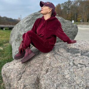 Izabela Kuligowska stylizka w bordowym welurowym dresie, czapce i rękawiczkach, siedząca na dużym głazie na tle parku, sportowa stylizacja jesienna.