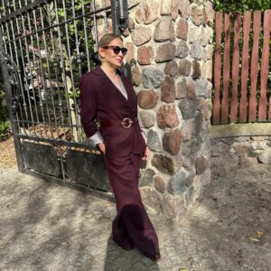 Izabela Kuligowska stylizka w burgundowym garniturze i okularach przeciwsłonecznych, oparta o metalową bramę przy kamiennym murze, jesienna stylizacja biznesowa.