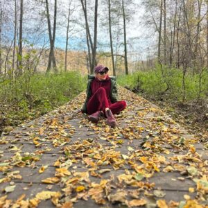 Izabela Kuligowska stylizka w sportowej stylizacji i kurtce, siedząca na drewnianym pomoście pokrytym jesiennymi liśćmi w lesie, modna turystyka i outdoor.