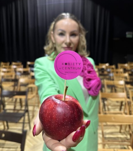 Izabela Kuligowska stylizka w jaskrawo zielonej marynarce, trzymająca jabłko z wtyczką "Kobiety w Centrum Mazowsze", scena, event motywacyjny.
