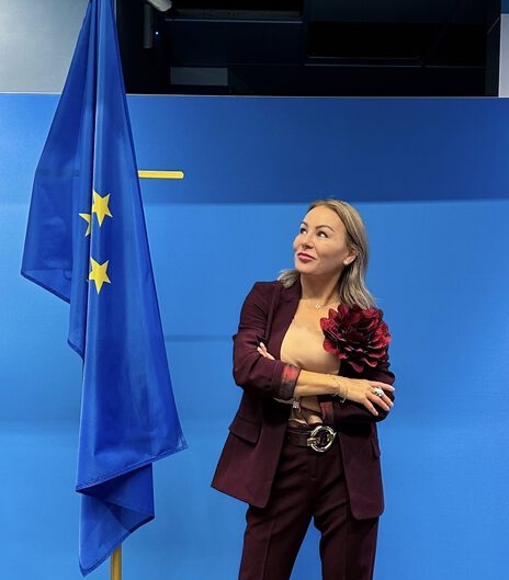 Izabela Kuligowska stylizka w bordowym garniturze ze spinką w kształcie kwiatu, stojąca przy fladze Unii Europejskiej na tle niebieskiej ściany, wizerunek biznesowy.