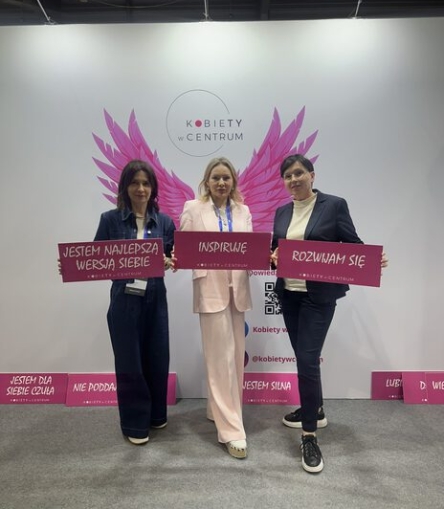 Izabela Kuligowska stylizka w różowym garniturze z dwiema innymi kobietami na tle stoiska "Kobiety w Centrum", trzymające hasła motywacyjne "Inspiruję", "Jestem najlepsza wersja siebie".