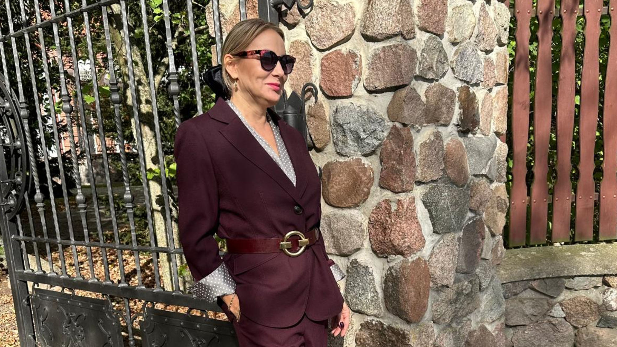 Izabela Kuligowska stylizka w burgundowym garniturze i okularach przeciwsłonecznych, oparta o metalową bramę przy kamiennym murze, jesienna stylizacja biznesowa.