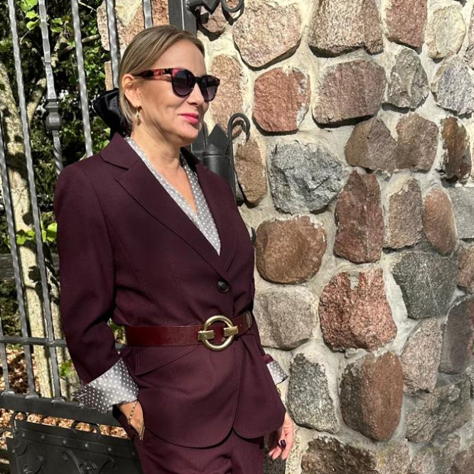 Izabela Kuligowska stylizka w burgundowym garniturze i okularach przeciwsłonecznych, oparta o metalową bramę przy kamiennym murze, jesienna stylizacja biznesowa.
